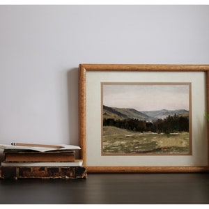 May include: A framed landscape painting of a valley with rolling hills and a forest in the distance. The painting is in a wooden frame with a light brown mat.