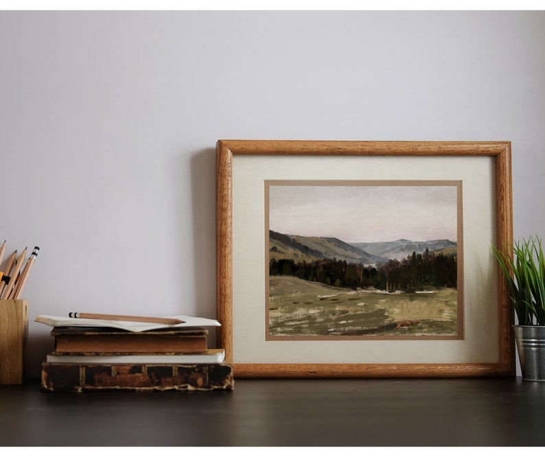 May include: A framed landscape painting of a valley with rolling hills and a forest in the distance. The painting is in a wooden frame with a light brown mat.