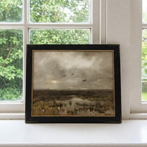 May include: A framed painting depicting a landscape with a cloudy sky and a flock of birds flying over a marsh. The painting is in a black frame with a gold inner border.