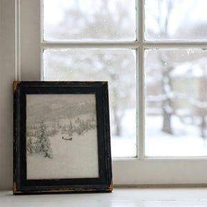 May include: A framed black and white landscape print of a snowy scene, resting on a white windowsill. The black frame has a distressed, aged appearance. The window behind the frame shows a snowy outdoor view.