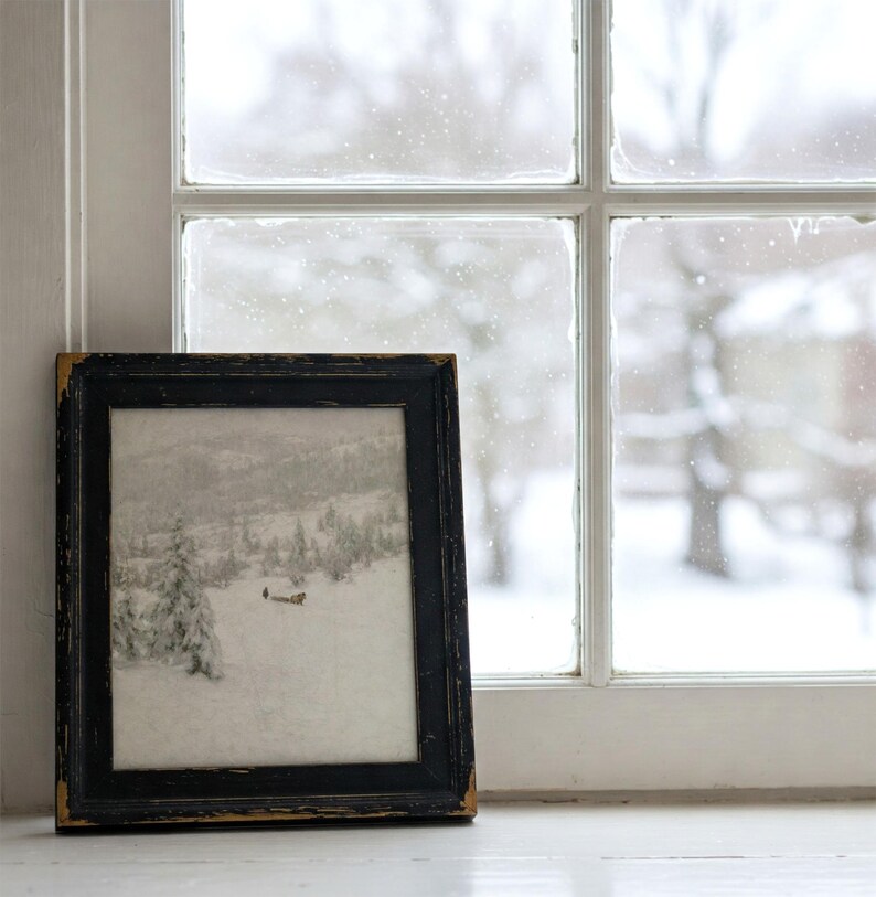 May include: A framed black and white landscape print of a snowy scene, resting on a white windowsill. The black frame has a distressed, aged appearance. The window behind the frame shows a snowy outdoor view.