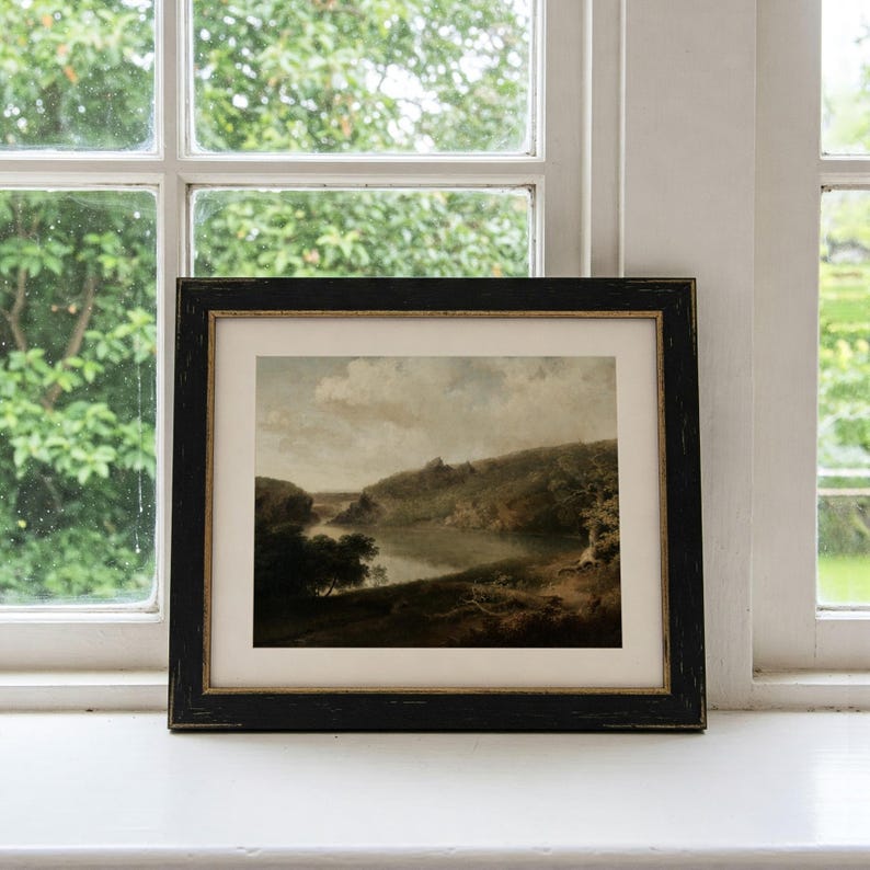 May include: A framed landscape painting depicting a river winding through a lush green valley. The painting is set against a cloudy sky and features a dark wooden frame.
