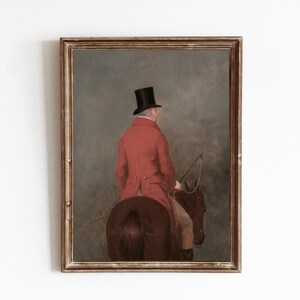 May include: A framed oil painting depicting a man in a red coat and black top hat riding a brown horse. The man is facing away from the viewer and holding a riding crop. The background is a hazy landscape.