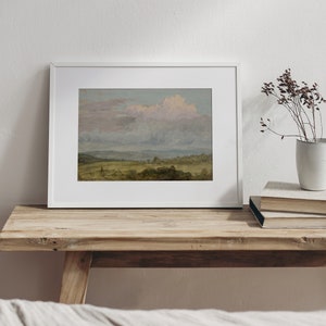 May include: A framed landscape painting depicting a field with a cloudy sky. The painting is in a white frame and is hanging on a wooden shelf.