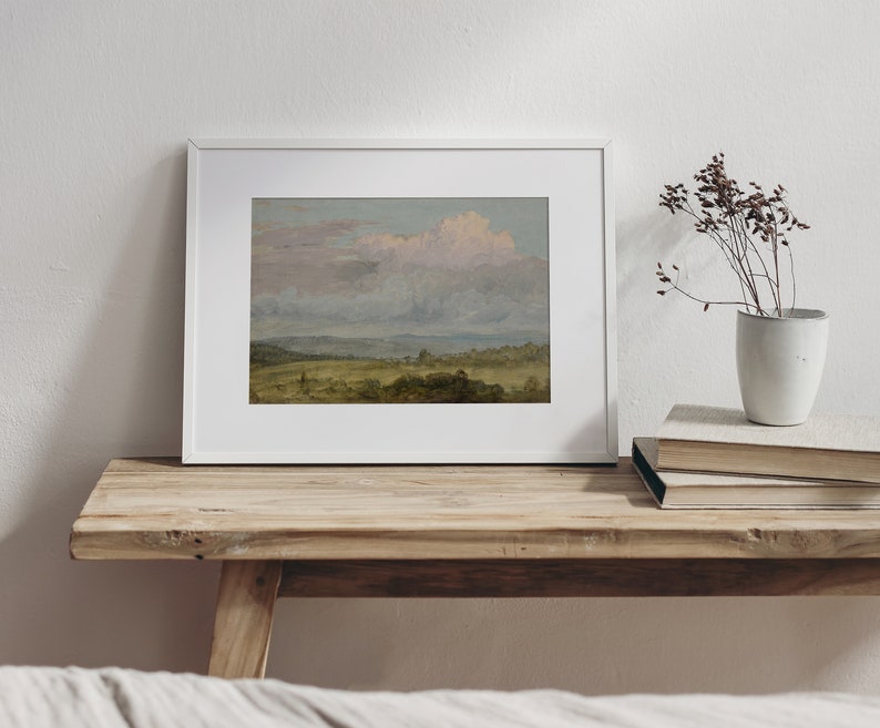 May include: A framed landscape painting depicting a field with a cloudy sky. The painting is in a white frame and is hanging on a wooden shelf.