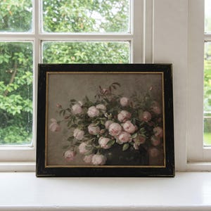 May include: A framed floral print featuring a bouquet of pink roses in a dark vase. The artwork is set against a muted background and framed in a black wooden frame with a gold inner edge. The print is displayed on a white windowsill.
