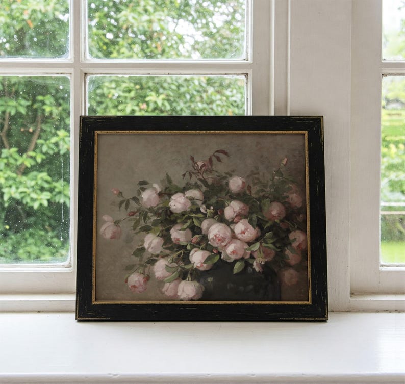 May include: A framed floral print featuring a bouquet of pink roses in a dark vase. The artwork is set against a muted background and framed in a black wooden frame with a gold inner edge. The print is displayed on a white windowsill.