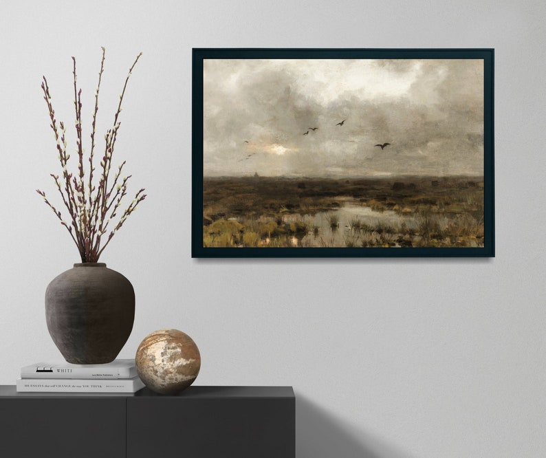 May include: A framed painting depicting a landscape with a cloudy sky and a flock of birds flying over a marsh. The painting is in a black frame and is hanging on a white wall.
