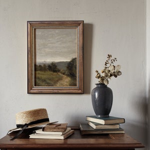 May include: A framed landscape painting of a rural scene with a path leading through a field, a vase with dried flowers, and a straw hat on a wooden table.