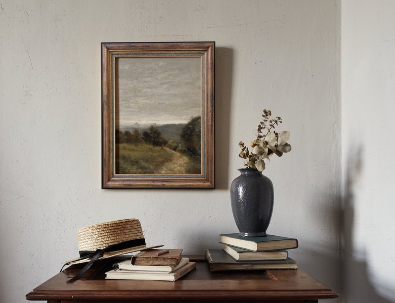 May include: A framed landscape painting of a rural scene with a path leading through a field, a vase with dried flowers, and a straw hat on a wooden table.