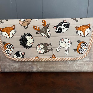 Op de afbeelding: Een grijze gewatteerde clutch met een bruine rand en een patroon van verschillende bosdieren, waaronder vossen, uilen, herten, wasberen, egels en konijnen.