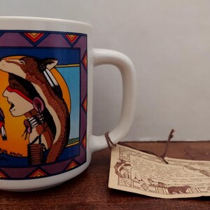 May include: White ceramic mug with a Native American design. The mug features a colorful illustration of Native American figures, a brown and white animal, and geometric patterns. A tag is attached to the handle.