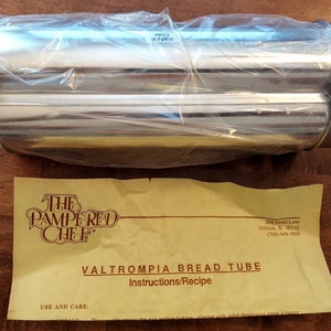 Puede incluir: Un tubo para pan de acero inoxidable, empaquetado en plástico transparente, con una hoja de instrucciones amarilla. La hoja dice "The Pampered Chef" y "VALTROMPIA BREAD TUBE Instructions/Recipe". El tubo es cilíndrico con interior acanalado.