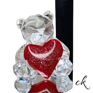 May include: A clear, faceted crystal bear figurine holding a red heart. A silver ring with a clear stone sits on a black display stand. The image has a white background.