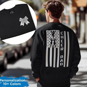 Op de afbeelding: Zwart sweatshirt met een wit Amerikaans vlagontwerp met een silhouet van cheerleaders en de tekst "CHEER DAD".