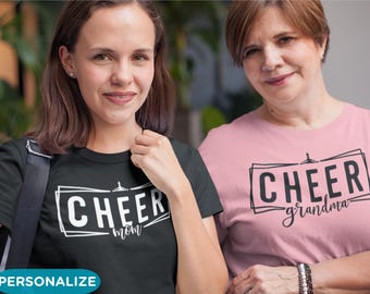 May include: Two women wearing black and pink t-shirts with the text "Cheer Mom" and "Cheer Grandma" printed on them. The shirts are a popular choice for cheerleading events.