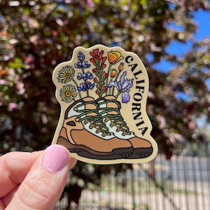 May include: A sticker of hiking boots with a bouquet of wildflowers growing out of them. The sticker is beige with brown outlines and the word "CALIFORNIA" is written vertically on the right side.