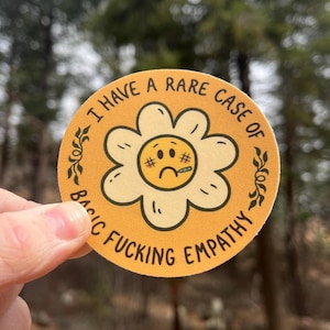 May include: A round sticker with a yellow background and a daisy design. The daisy has a sad face with a thermometer and hashtags. The text reads "I HAVE A RARE CASE OF BASIC FUCKING EMPATHY" in a circular pattern around the daisy.