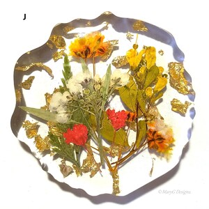 May include: A round, clear resin coaster with a pressed flower design. The coaster features a variety of flowers in shades of red, white, yellow, and green, with gold leaf accents. The coaster is approximately 4 inches in diameter.