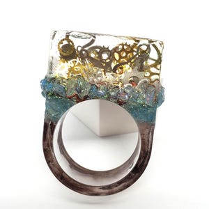 May include: A ring featuring a rectangular clear resin top embedded with various metallic clockwork gears. The base of the resin section is adorned with a band of iridescent blue crushed stone fragments, while the ring band is a dark, translucent material.