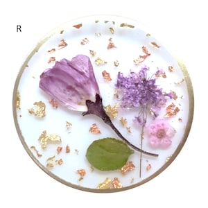 May include: A round, white resin coaster with a gold rim. The coaster features pressed flowers, including a purple flower, a cluster of small purple flowers, and a pink flower. The coaster is decorated with gold and copper flakes.