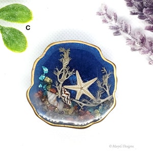 Op de afbeelding: Een blauwe hars-briefgewicht met een gouden rand, met een witte zeester, schelpen en koraal. Het briefgewicht is gevuld met glitter en andere kleine decoratieve elementen.