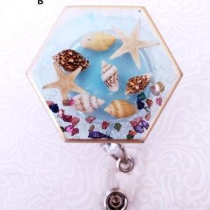 Puede incluir: Un portagafete hexagonal de resina con fondo azul y varias conchas marinas y una estrella de mar. El portagafete está unido a un carrete retráctil de gafete plateado.