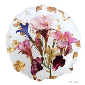 May include: A round, white resin coaster with embedded dried flowers in shades of pink, purple, and yellow. Gold leaf accents are scattered throughout the coaster, adding a touch of elegance. The coaster has a wavy edge.