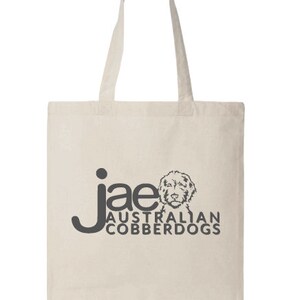 Puede incluir: Bolso tote de color crema con asas largas. El bolso presenta el texto "jae AUSTRALIAN COBBERDOGS" en gris oscuro, junto con un gr&aacute;fico de la cara de un perro. El bolso est&aacute; hecho de una tela resistente.