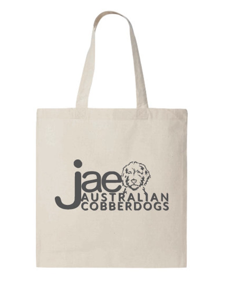 Puede incluir: Bolso tote de color crema con asas largas. El bolso presenta el texto "jae AUSTRALIAN COBBERDOGS" en gris oscuro, junto con un gr&aacute;fico de la cara de un perro. El bolso est&aacute; hecho de una tela resistente.