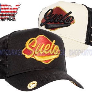 May include: Two black and white trucker hats with a red and gold embroidered logo that says "Suelo" on the front. The hats have a mesh back and a black adjustable strap.