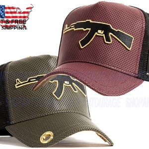 May include: Two trucker hats with a black mesh back and a perforated leather front. The front of the hats feature a gold and black graphic of an AK-47 assault rifle. One hat is burgundy and the other is olive green.