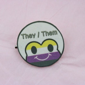 Puede incluir: Un pin redondo con un diseño de cara de rana, con los colores de la bandera del orgullo no binario: amarillo, blanco, morado y negro. El pin tiene las palabras "They / Them". El pin está sobre un fondo rosa.