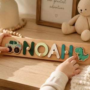 May include: Wooden name puzzle spelling "NOAH" with a green monster truck and a green dinosaur. The letters are in various shades of green and white. The puzzle is on a light wood base.