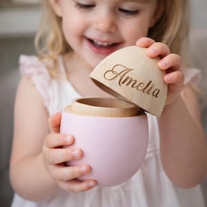 May include: A pink wooden keepsake box with a natural wood lid, engraved with the name "Amelia". The box is held open, showing the interior. The box is a soft pink colour.
