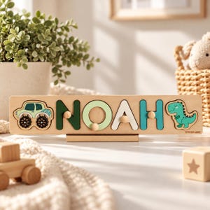 May include: A wooden name puzzle board featuring the name NOAH in cutout letters with small round knobs. The board includes a green monster truck on the left and a green dinosaur on the right. The text TOYS FOR LOGIC is printed on the right.