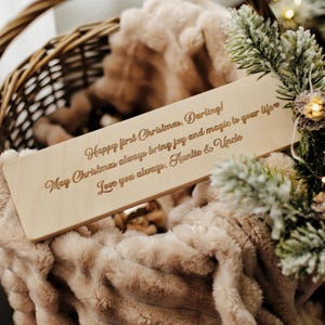 May include: A wooden rectangular sign with engraved text, resting on a plush, beige blanket inside a wicker basket. The sign reads "Happy first Christmas, Darling! May Christmas always bring joy and magic to your life. Love you always. Auntie & Uncle."