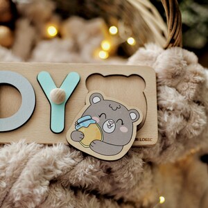 May include: Wooden puzzle with the letters "D", "Y", and a picture of a bear holding a jar of honey. The puzzle is on a light brown surface.