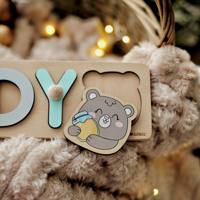 May include: Wooden puzzle with the letters "D", "Y", and a picture of a bear holding a jar of honey. The puzzle is on a light brown surface.