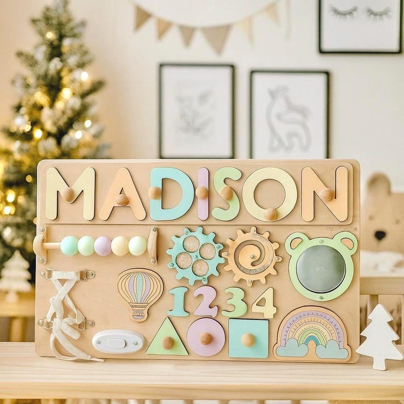 Op de afbeelding: Een houten activiteitenbord met de naam "MADISON" in pastelkleuren. Het bord heeft verschillende interactieve elementen, waaronder tandwielen, vormen, cijfers, een spiegel en een regenboog. Een leuk en leerzaam speelgoed voor peuters.