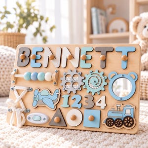 May include: A wooden sensory activity board featuring the name BENNETT in blue, white, and gray letters. The board includes a bead maze, spinning gears with small white balls, a mirror, a lace-up shoe detail, numbers 1234, and shapes.