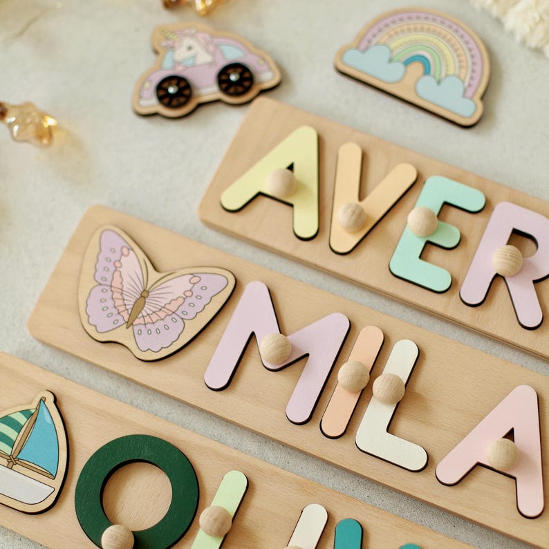 May include: Wooden name puzzles with pastel-coloured letters and wooden pegs. The puzzles feature the names "AVER" and "MILA". Decorative wooden shapes include a butterfly, a rainbow, a car with a unicorn, and a sailboat.