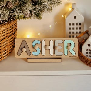 May include: A wooden name puzzle with the name "ASHER" spelled out in colorful letters. The puzzle is on a wooden stand and has small wooden knobs for each letter.
