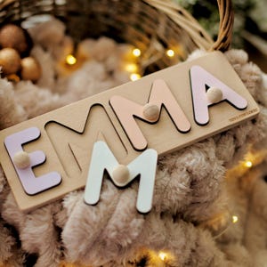 May include: Wooden name puzzle spelling "EMMA" with pastel-colored letters and wooden knobs. The letters are light pink, lavender, and white, set on a light wood base. The puzzle is designed for children and features the text "TOYS FOR LOGIC".