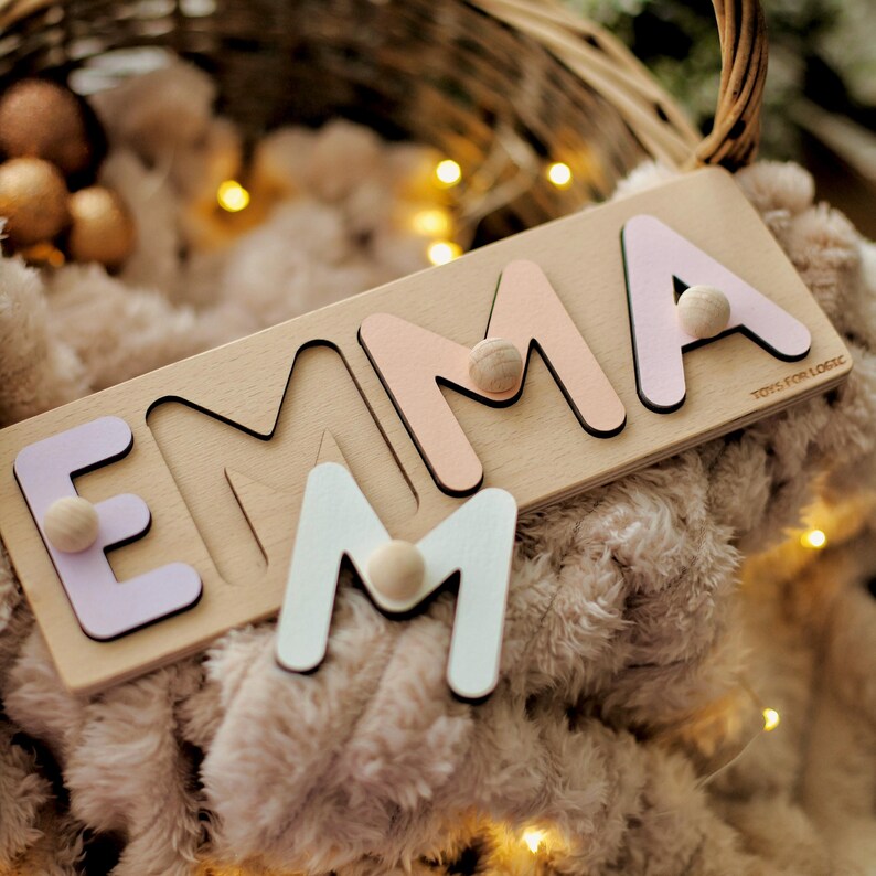 May include: Wooden name puzzle spelling "EMMA" with pastel-colored letters and wooden knobs. The letters are light pink, lavender, and white, set on a light wood base. The puzzle is designed for children and features the text "TOYS FOR LOGIC".