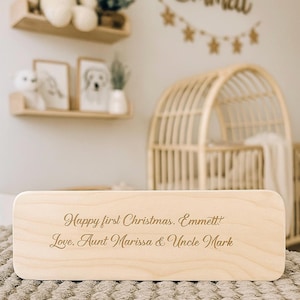 May include: A light-coloured wooden box with rounded edges. The box has the inscription "Happy first Christmas. Emmett Love, Aunt Marissa & Uncle Mark" in a cursive font. The box is sitting on a textured surface.