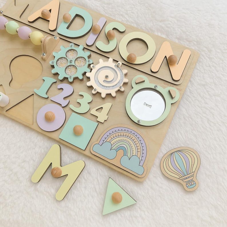 Op de afbeelding: Houten educatief puzzelbord met de naam "ADISON" in pastelkleuren. Inclusief vormen, cijfers, tandwielen, een spiegel en een luchtballon. De puzzelstukken hebben houten knoppen voor eenvoudige bediening. Een leuk en boeiend leerspeelgoed.