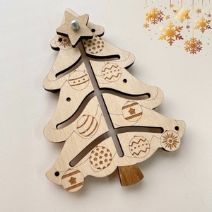 Puede incluir: Un adorno de &aacute;rbol de Navidad de madera con una estrella en la parte superior y recortes para adornos. El adorno es de color marr&oacute;n claro y tiene un acabado de madera natural.