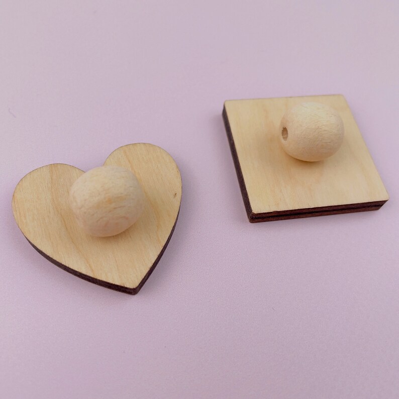 Puede incluir: Dos formas de madera, un coraz&oacute;n y un cuadrado, cada una con una cuenta redonda de madera en el centro.