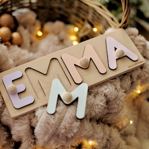 May include: A wooden puzzle spelling "EMMA" with pastel-coloured letters and wooden knobs. The letters are light pink, lavender, and white. The puzzle is on a light-coloured wooden base. The text "TOYS FOR LOGIC" is printed on the puzzle.